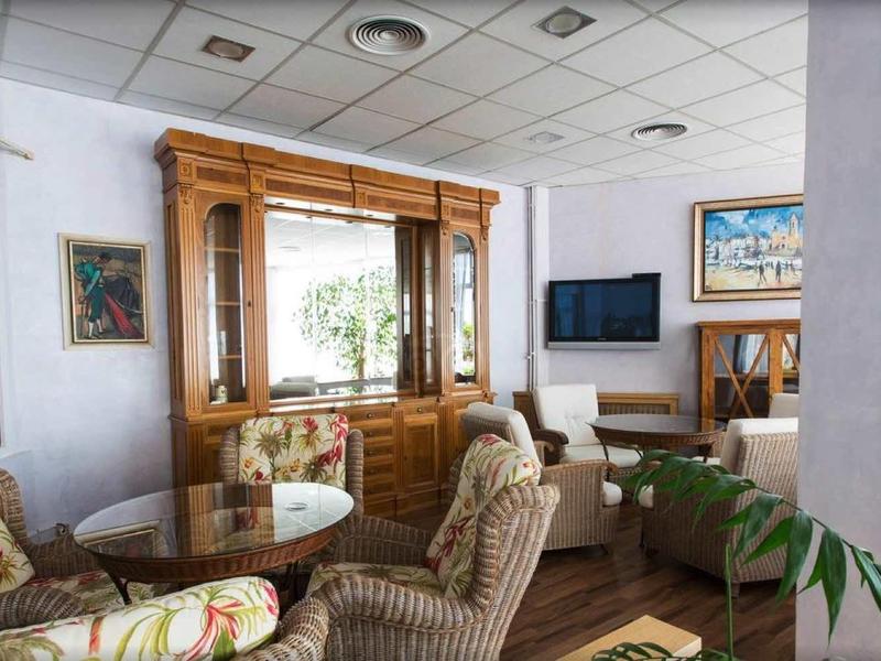 Cozy hotel lounge with wooden windows, cushioned chairs, plants, and a TV on the wall.
