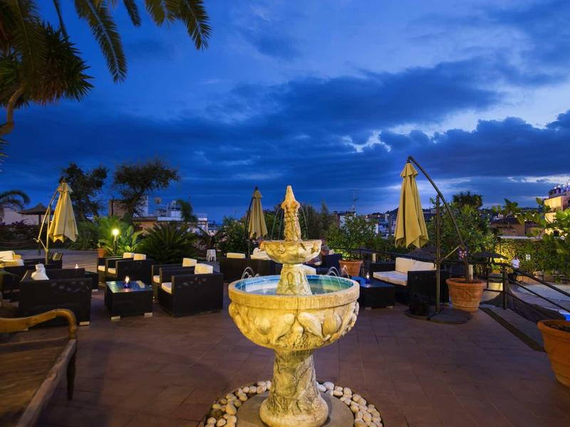 Outdoor hotel terrace at dusk with a central stone fountain, plants, and umbrellas under a cloudy sky.