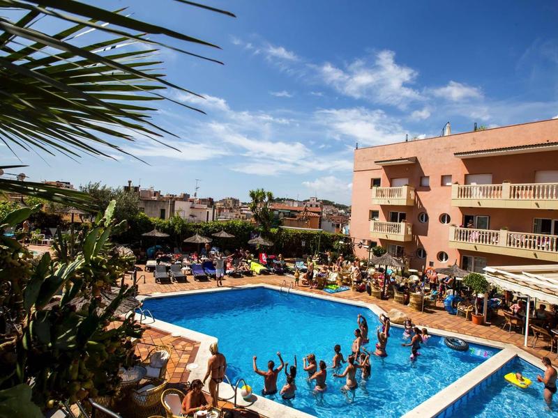 Outdoor hotel pool with people swimming and lounging under a sunny sky with palm trees.
