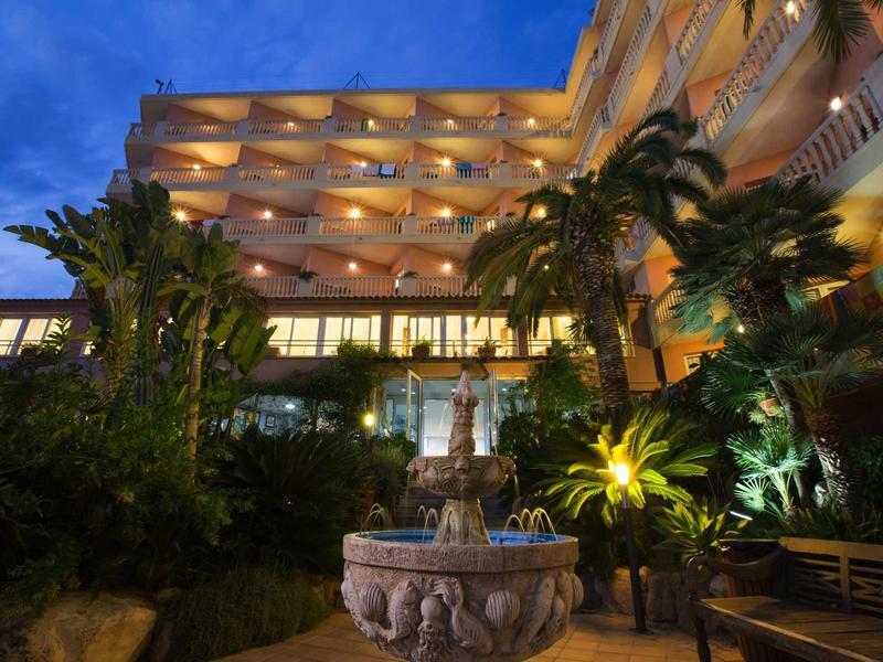 Hotel exterior at dusk with balconies, palm trees, and a decorative fountain in the garden area.