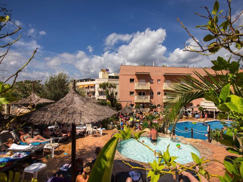 Outdoor hotel pool area with sun loungers, umbrellas, and a pink building under a blue sky.