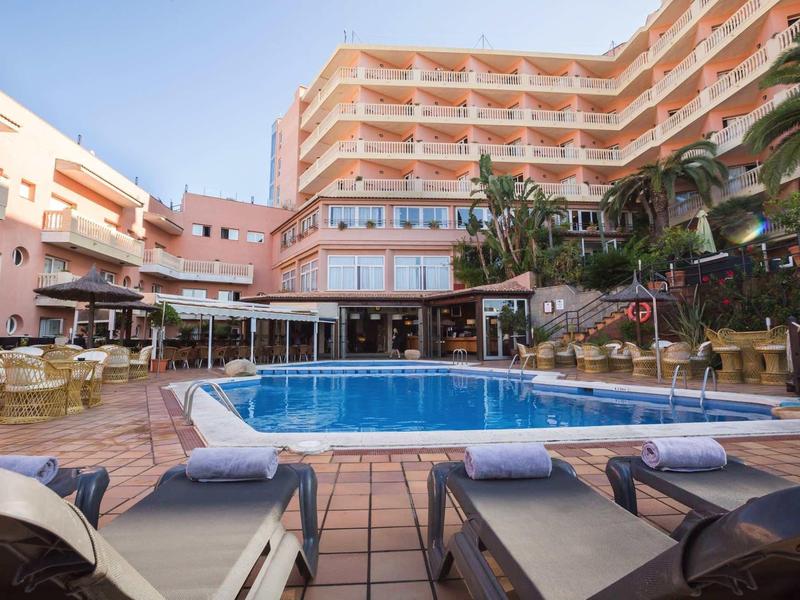 Hotel pool area with loungers, towels, umbrellas, and a multi-story building under clear sky.