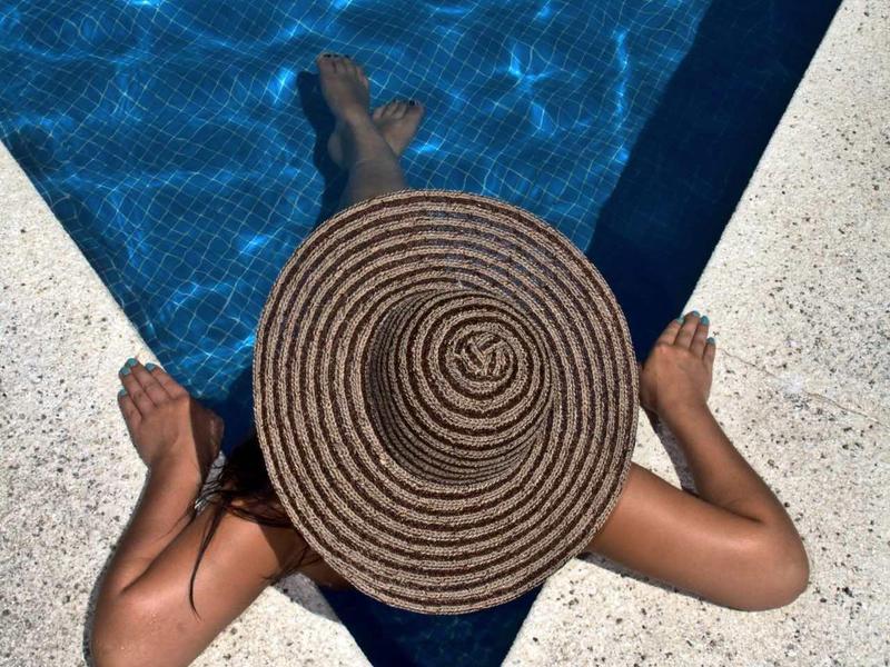 Person relaxing at the edge of a pool wearing a large striped sunhat.