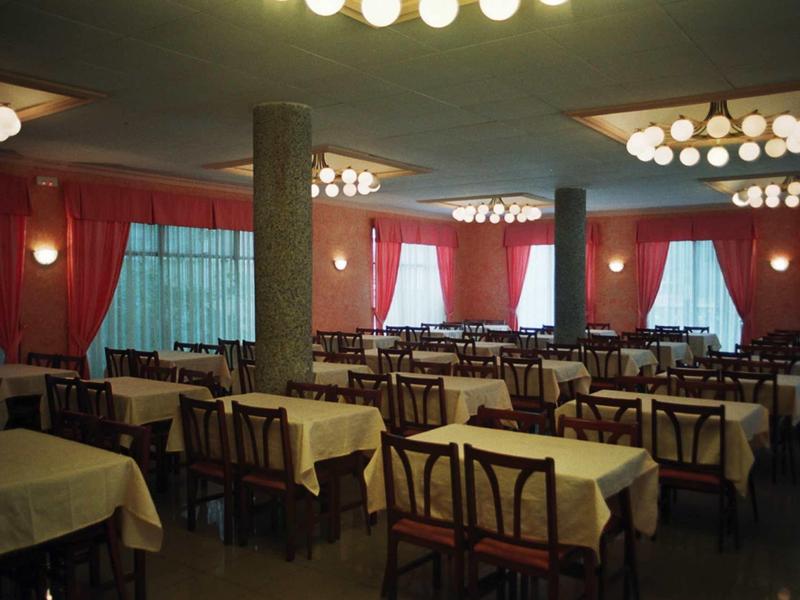 Duża jadalnia z wieloma stołami i krzesłami, czerwonymi zasłonami i okrągłymi lampami sufitowymi.