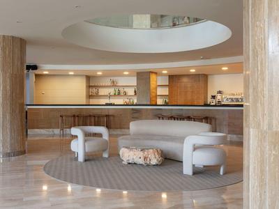 Moderner Hotel-Lobbybereich mit runder grauer Couch, Teppich und Bar im Hintergrund, helle Marmorsäulen.