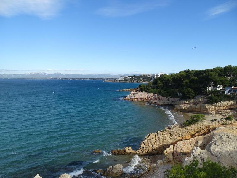 Küste mit blauem Meer, felsigem Ufer und grünem Wald unter blauem Himmel mit wenigen Wolken.