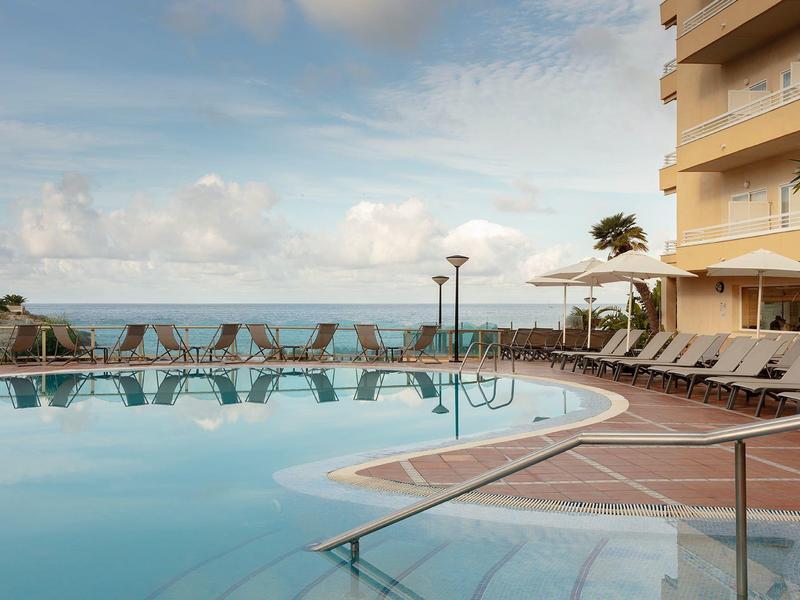 Großer, klarer Pool mit vielen Liegestühlen am Rand, Palmen, Gebäude und Meer im Hintergrund.