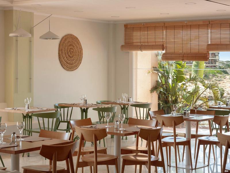 Moderner heller Gastraum mit Holzstühlen, weißen Tischen und großem Fenster mit Pflanzen draußen.