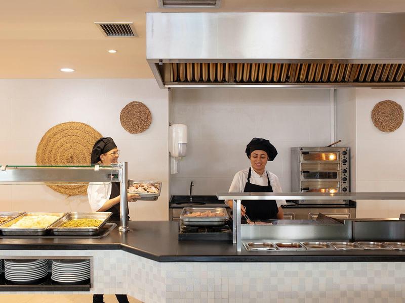 Zwei Köchinnen in weißer Kleidung und schwarzen Hauben arbeiten in einer modernen, sauberen Kantine.