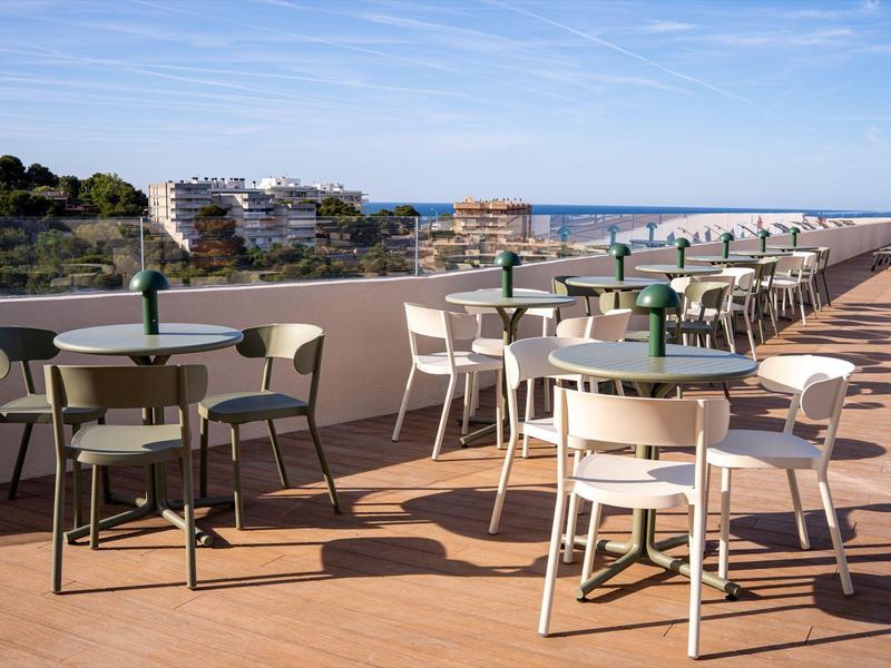 Leere Außenterrasse mit Tischen und Stühlen, Meer und Gebäude im Hintergrund.