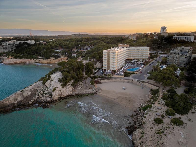 Hotel an einer Küste mit Strand, Klippen und bewaldeten Hügeln bei Sonnenuntergang.