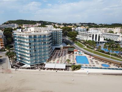 Nowoczesny budynek hotelowy z basenem i leżakami na plaży pod bezchmurnym niebem.