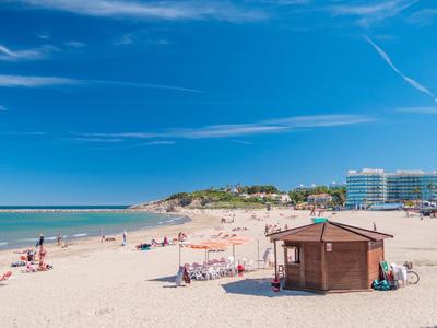 Szeroka piaszczysta plaża z leżakami, drewnianą chatką i hotelami w tle pod niebieskim niebem.
