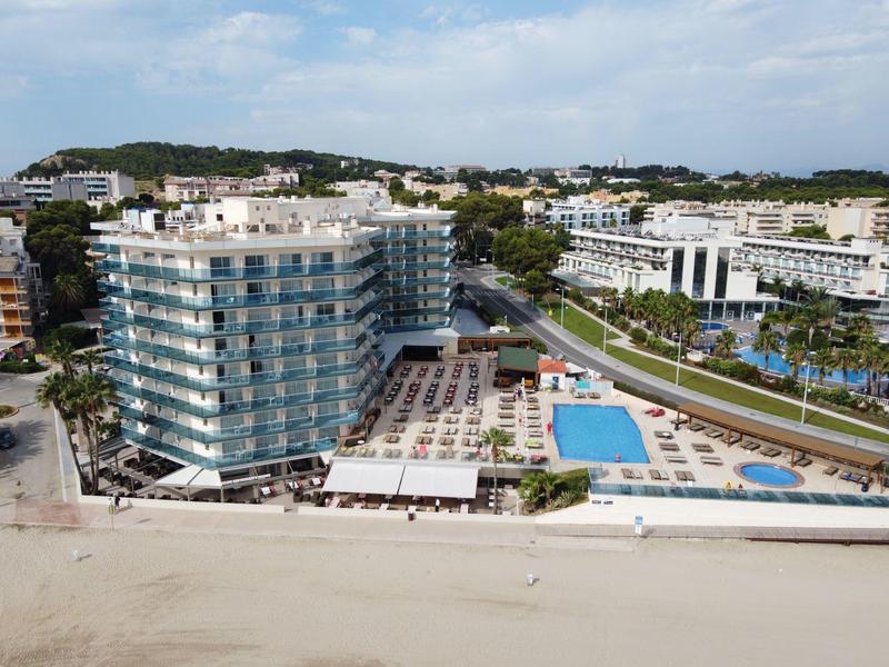Nowoczesny budynek hotelowy z basenem i leżakami na plaży pod bezchmurnym niebem.