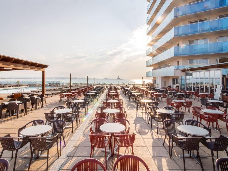 Duży taras z wieloma stolikami i krzesłami obok nowoczesnego budynku hotelowego z widokiem na morze.