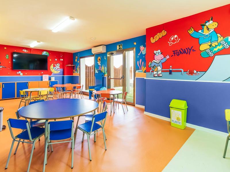 Kolorowy pokój zabaw dla dzieci z stołami, krzesłami i muralami na ścianach w kolorze czerwonym i niebieskim.