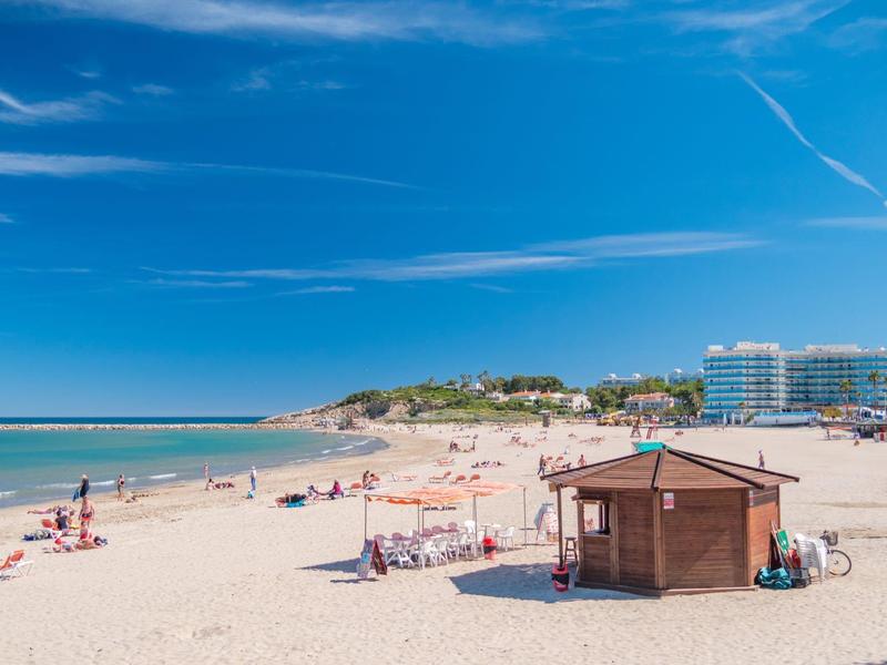 Szeroka piaszczysta plaża z leżakami, drewnianą chatką i hotelami w tle pod niebieskim niebem.