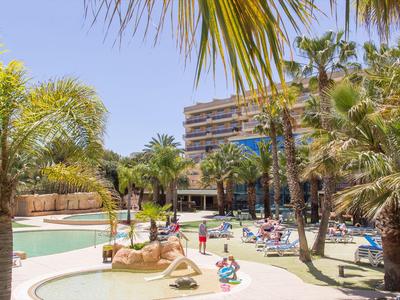 Hotelpool mit bunten Kinderwasserfontänen und Palmen unter blauem Himmel.