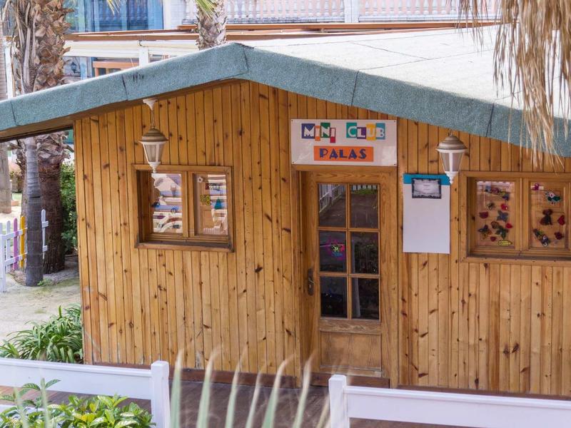 Small wooden cabin with windows and a door, surrounded by plants and wooden fence panels.