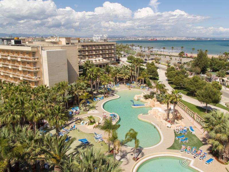 Resort hotel with curved pool, sun loungers, palm trees, and ocean view under a partly cloudy sky.