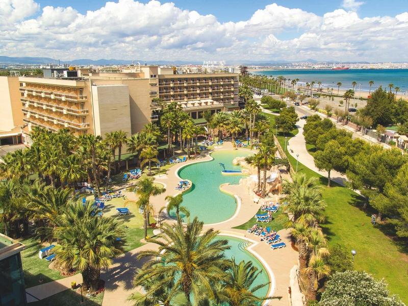 Resort hotel with pool, palm trees, and beachside view under a blue sky with clouds.