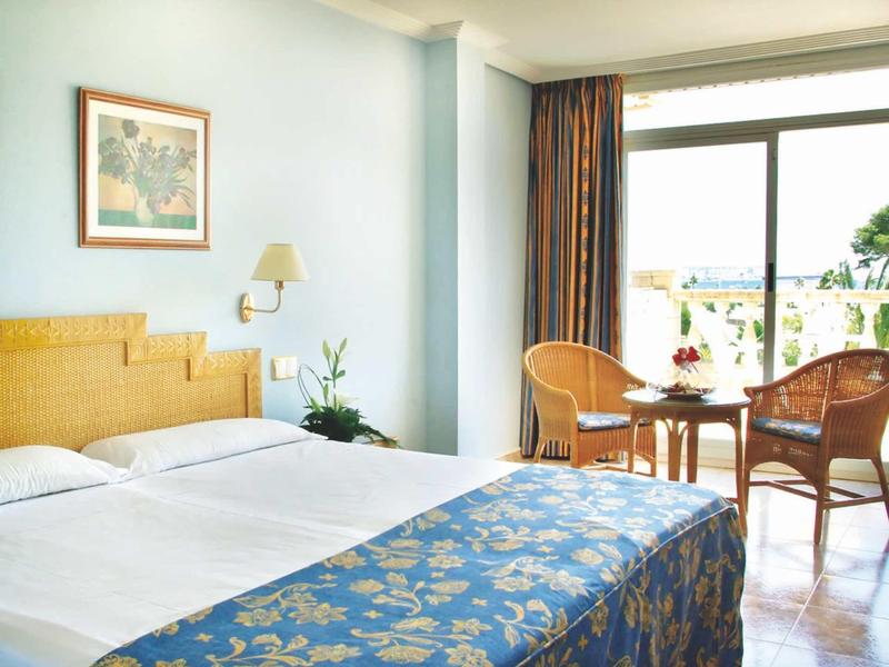 Bright hotel room with a double bed, floral bedspread, and a balcony with chairs and a table.