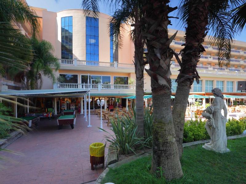 Hotel exterior with palm trees, garden, and outdoor seating area beneath a canopy.