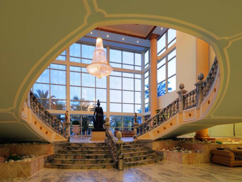 Spacious hotel lobby with large windows, elegant chandelier, curved staircase, and seating areas.