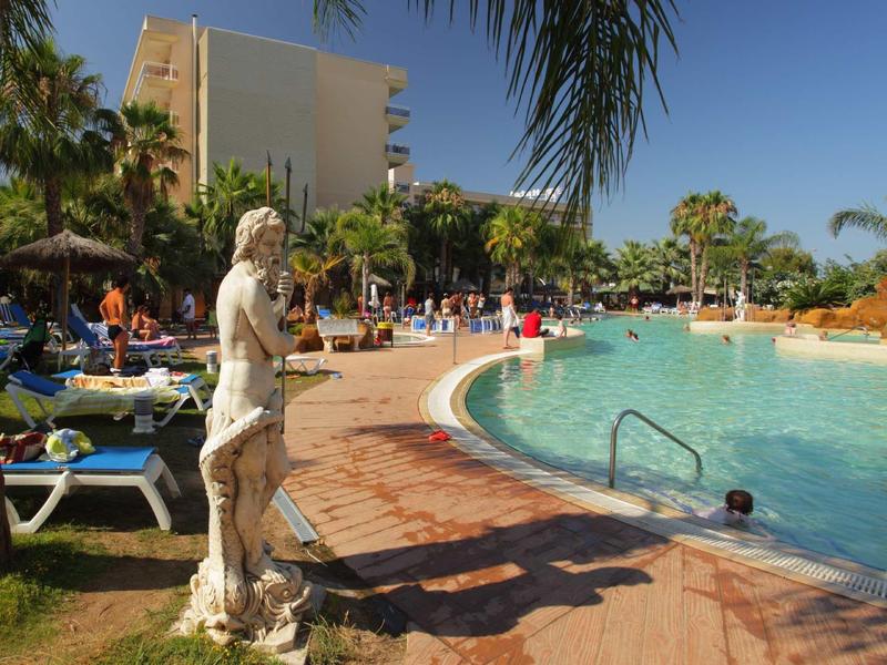 Sunny hotel pool area with statue, lounge chairs, palm trees, and blue sky