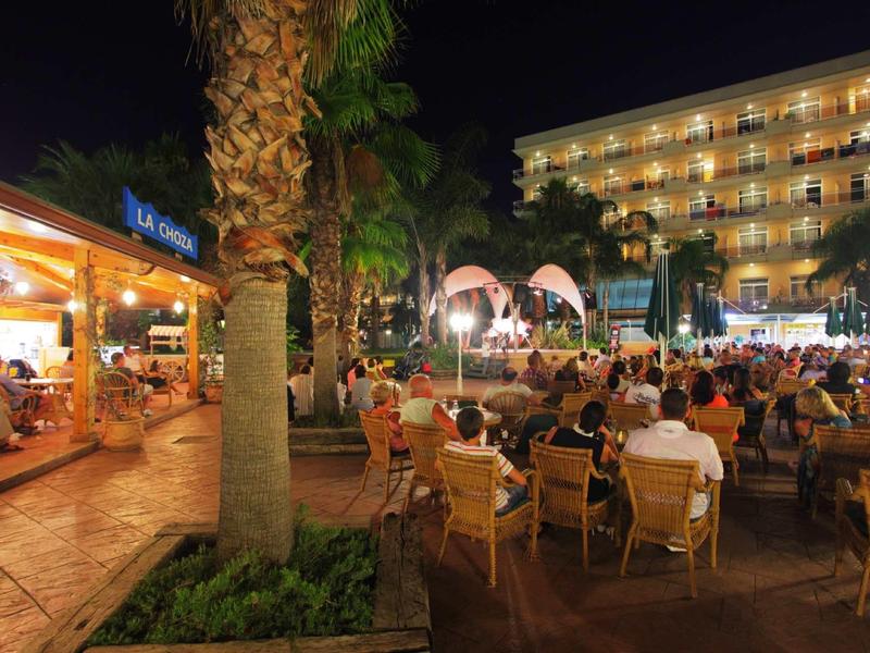 Outdoor night seating area at a lit hotel with palm trees and guests dining and socializing.