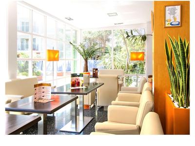 Heller Warteraum mit beigen Sesseln, Glasfenstern, grünen Pflanzen und orangenen Wänden.