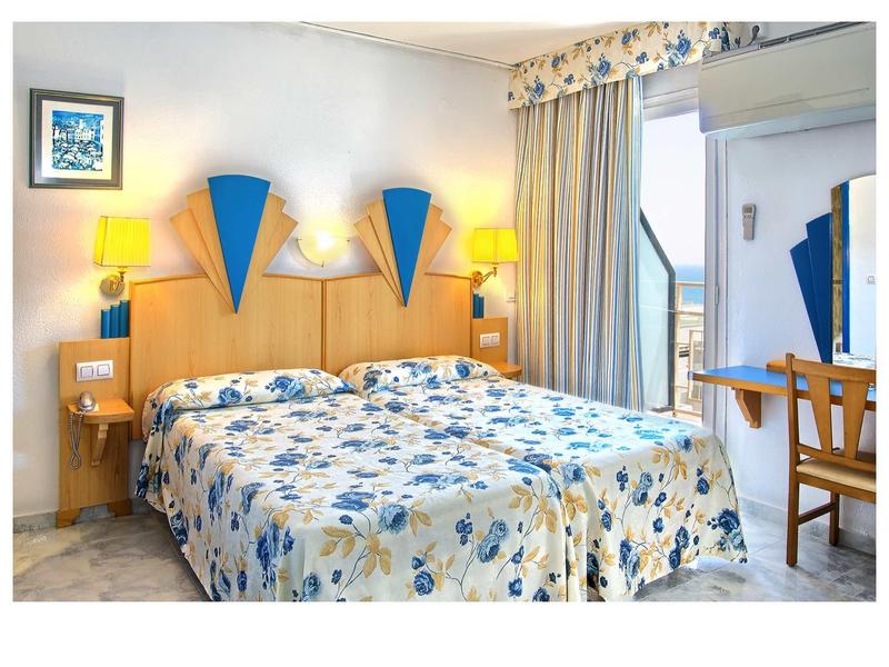 Hell eingerichtetes Hotelzimmer mit Doppelbett, floraler Bettwäsche und Holzmöbeln.