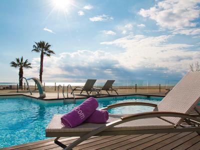 Piscina soleggiata con lettini, asciugamani viola, palme e vista sul mare sotto un cielo azzurro.
