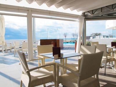 Terrazza aperta e luminosa dell'hotel con sedie beige, tavoli in vetro e vista mare sotto un tetto a lamelle.