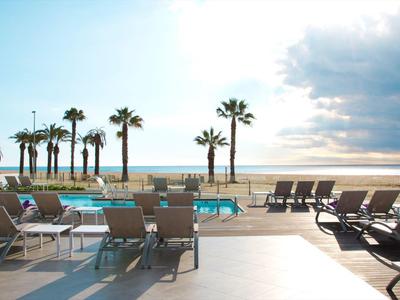 Poolbereich mit Liegestühlen, Palmen vor dem Strand und bewölktem Himmel im Hintergrund.