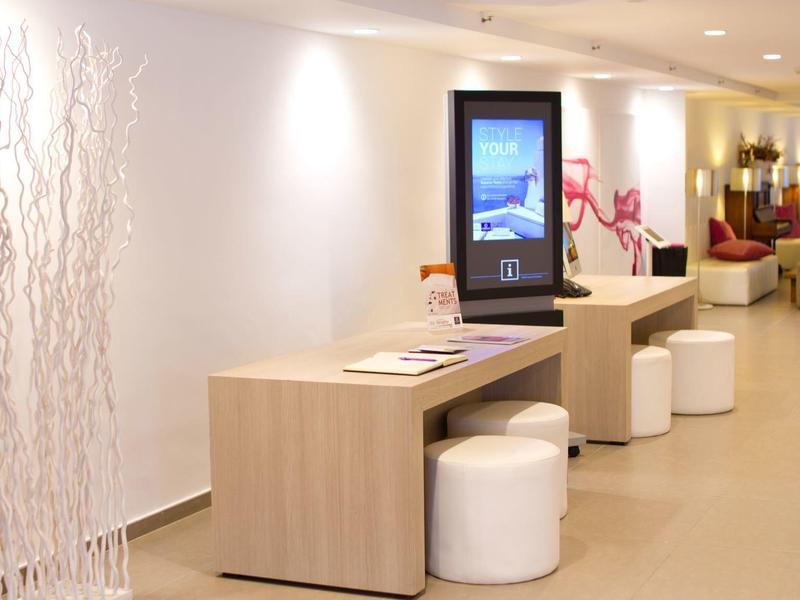 Moderna hall dell'hotel con tavoli in legno chiaro, sgabelli bianchi e chiosco informativo digitale.
