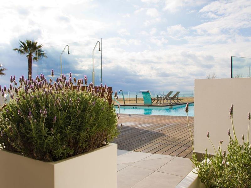 Zona piscina all'aperto con lettini, palme e piante di lavanda sotto un cielo nuvoloso.