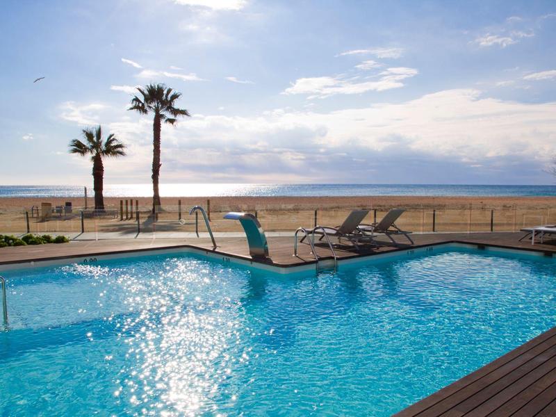 Piscina all'aperto con lettini e palme vicino alla spiaggia sotto un cielo limpido.