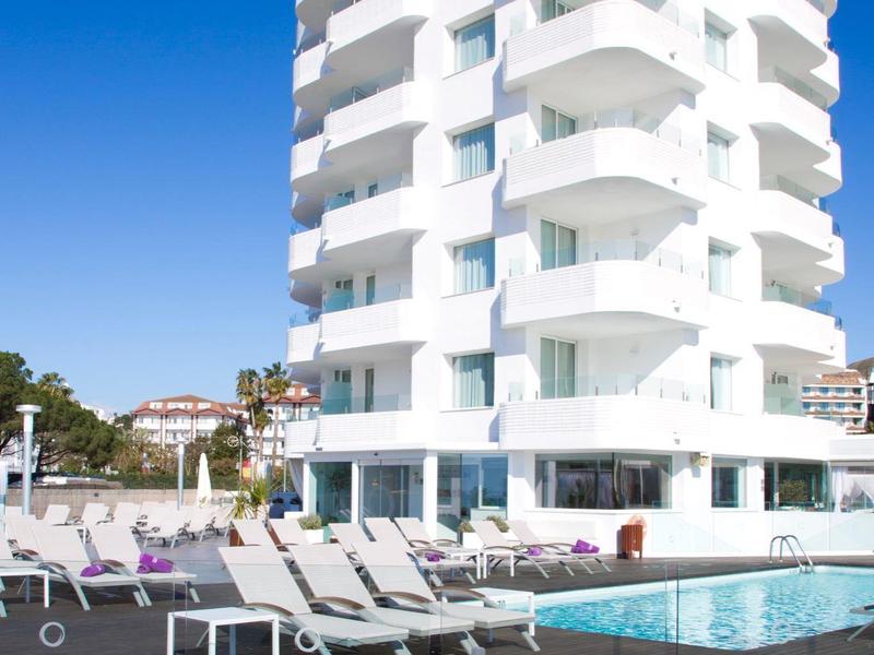 Hotel bianco su più piani con balconi affacciati su piscina all'aperto e lettini.