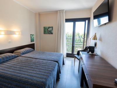 Habitación de hotel luminosa con dos camas individuales, escritorio, televisor y balcón con vistas.