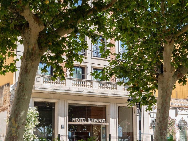 Vista de la fachada de un hotel detrás de dos grandes árboles con follaje verde denso.