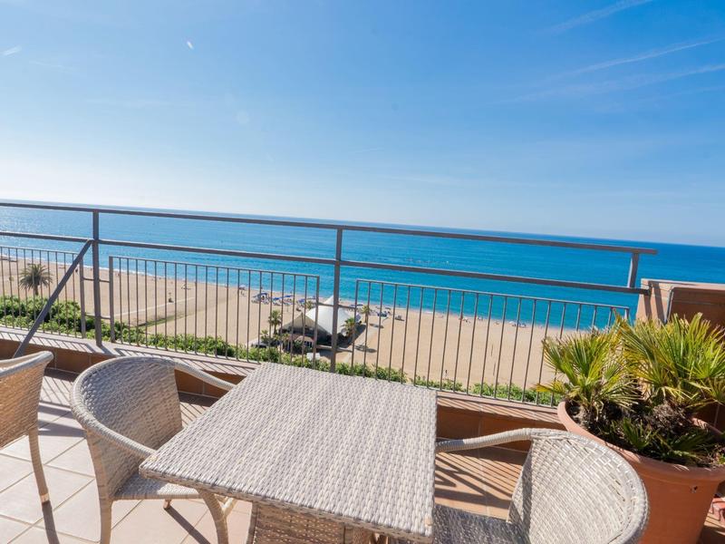 Terraza con mesa y sillas junto a una planta con vista al océano y la playa bajo cielo despejado.