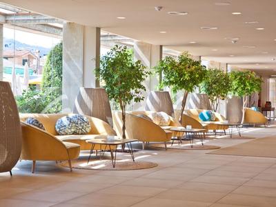 Heller Hotellobby-Bereich mit beige Sesseln, Tischen und grünen Pflanzen entlang der Fenster.