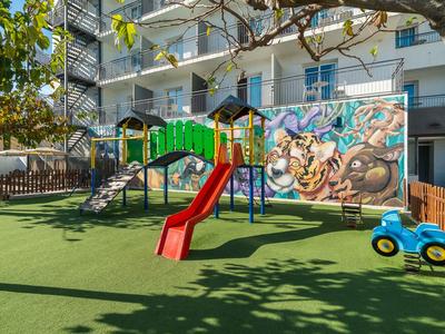 Kinder-Spielplatz mit Rutsche und Klettergerüst neben einem Hotel mit buntem Wandgemälde.