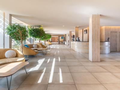 Helle Hotellobby mit Sitzbereichen, großen Fenstern, Pflanzen und Empfangstresen aus Holz.