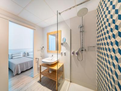 Helles Badezimmer mit offener Dusche und Blick auf ein Zimmer mit Bett.