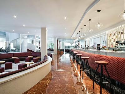 Moderner Hotelbar-Bereich mit roten Hockern und Sitzgelegenheiten, hellem Interieur und Marmorboden.