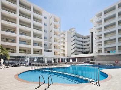 Outdoor swimming pool surrounded by white multi-story hotel buildings under a clear sky.