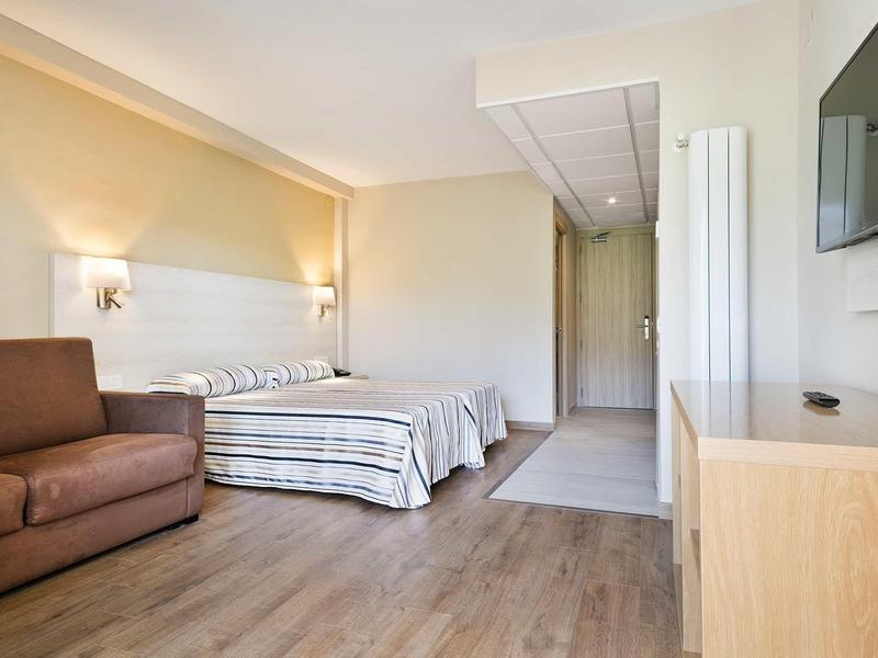 Bright hotel room with twin beds, sofa, wooden floor, and a wall-mounted TV.
