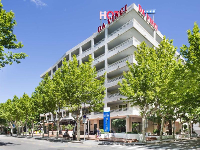 Modern hotel building with balconies, surrounded by trees and clear blue sky.